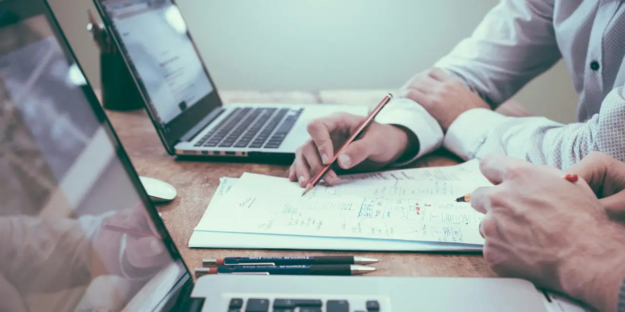 Dos personas trabajando con portátiles y un cuaderno de notas sobre una mesa
